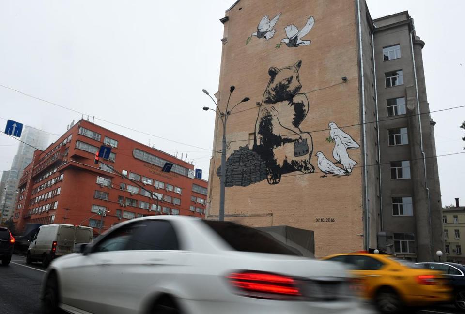 Cars driving past a bulding with graffiti showing a brown bear distributing bulletproof vests to doves of peace in central Moscow.